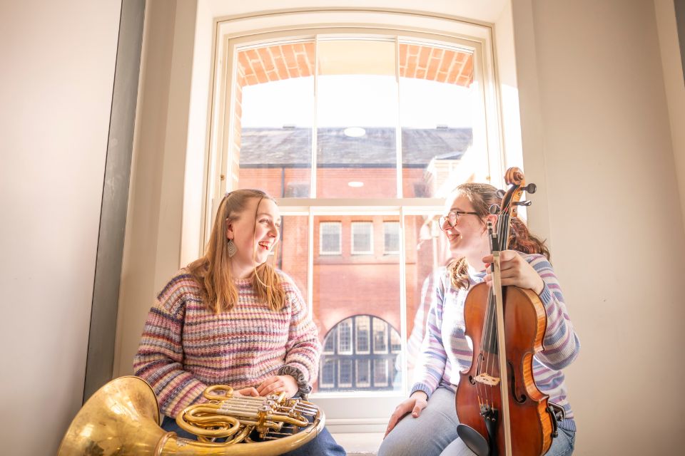 Two young white women sat in a sunny window frame. The woman on the left has a French Horn on her knee, the woman on the right is holding a viola